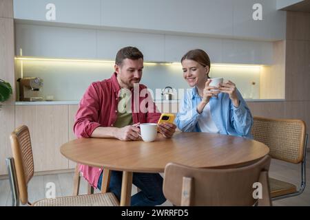 Happy fun man sitting in kitchen table with coffee talking showing smartphone to interested woman. Stock Photo