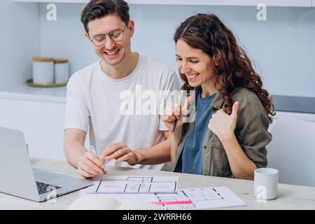 A smiling couple is working on a new apartment project, with a laptop and documents with drawings of the house spread out on the table. Family housewarming. Stock Photo