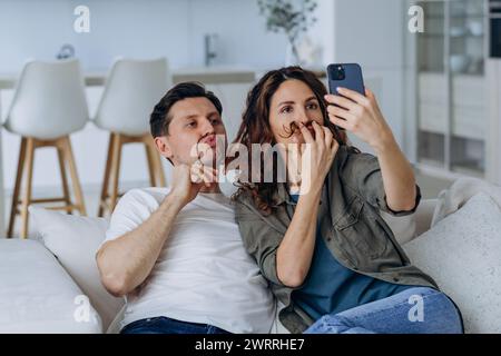 A playful couple captures a humorous selfie moment on the couch, with the woman making a fake mustache with her hair. Their spontaneous fun brings life to the relaxed home setting. Playful Couple Taking Selfies Stock Photo