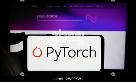 Person holding cellphone with logo of machine learning library PyTorch in front of webpage. Focus on phone display. Stock Photo