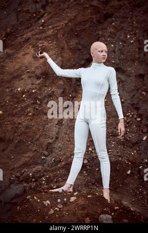Full length portrait of young hairless girl with alopecia in white futuristic suit holding handful of barren soil thoughtfully, highlighting potential Stock Photo