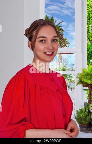 The photo shows the upper body of a young woman in a bright red blouse sitting in front of a window, smiling cheerfully. The natural light emphasizes Stock Photo