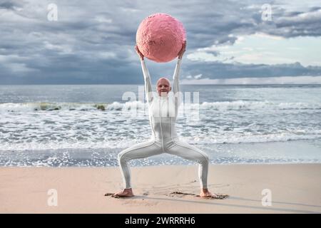 Hairless performer girl with alopecia in white futuristic suit dancing with pink sphere at sea, metaphoric surreal scene exudes confidence, unique bea Stock Photo