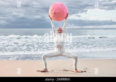 Hairless performer girl with alopecia in white futuristic suit dancing with pink sphere at sea, metaphoric surreal scene exudes confidence, unique bea Stock Photo