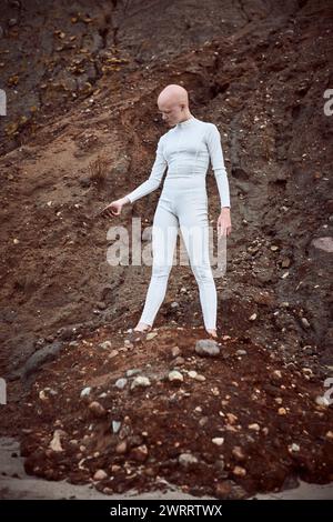 Full length portrait of young hairless girl with alopecia in white futuristic suit holding handful of barren soil thoughtfully, highlighting potential Stock Photo