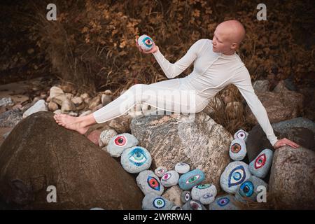 Young hairless girl with alopecia in white futuristic costume pensively examines stone with eye at surreal landscape, performance symbolizes introspec Stock Photo