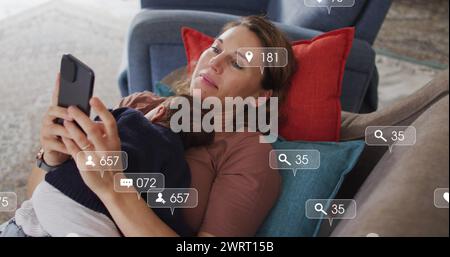 Image of social media icons with numbers over caucasian woman holding baby and using smartphone Stock Photo