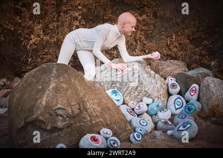 Young hairless girl with alopecia in white futuristic costume pensively examines stone with eye at surreal landscape, symbolizes introspection and ree Stock Photo