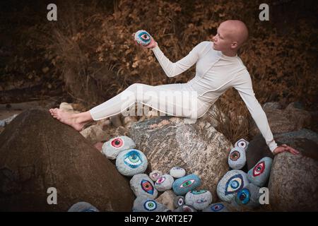 Young hairless girl with alopecia in white futuristic costume pensively examines stone with eye at surreal landscape, performance symbolizes introspec Stock Photo