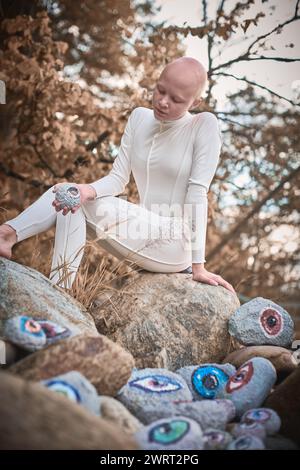 Young hairless girl with alopecia in white futuristic costume pensively examines stone with eye at surreal landscape, performance symbolizes introspec Stock Photo