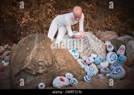 Young hairless girl with alopecia in white futuristic costume pensively examines stone with eye at surreal landscape, symbolizes introspection and ree Stock Photo