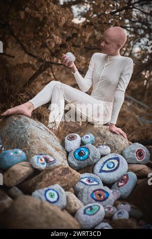 Young hairless girl with alopecia in white futuristic costume pensively examines stone with eye at surreal landscape, performance symbolizes introspec Stock Photo