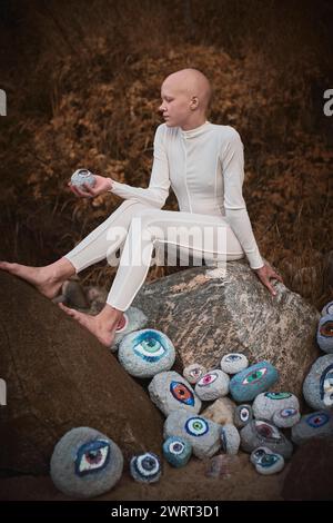 Young hairless girl with alopecia in white futuristic costume pensively examines stone with eye at surreal landscape, performance symbolizes introspec Stock Photo