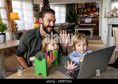 Happy single father with children waving hands while doing video call through laptop at home Stock Photo