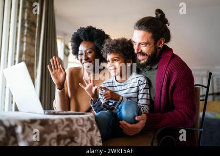 Happy modern multiethnic diverse parents and kid waving hands while using laptop for video chat Stock Photo