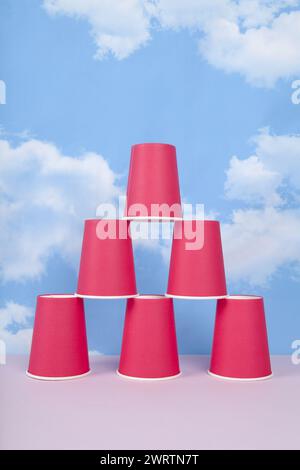 red paper cups mounted in a pyramid symbolizing success, balance and stability, in front of a blue summer sky with white clouds. Minimal still life ph Stock Photo