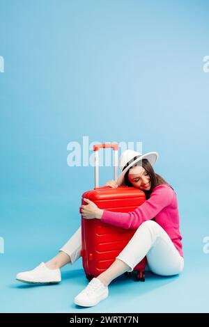 Happy young tourist woman sitting at the suitcase, hugging it and going to travel on holidays isolated on blue background Stock Photo