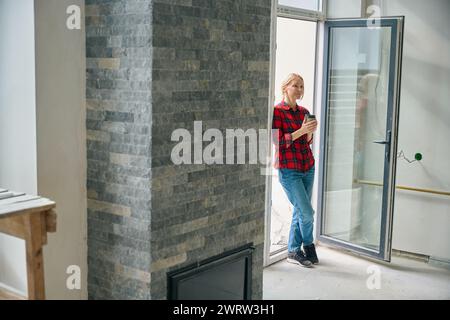 Full length photo of satisfied lady standing in doorway and holding paper glass in hands during coffee break Stock Photo