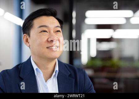 Dreamy and smiling man, Asian businessman sitting in the office in a suit and thoughtfully looking to the side, resting. Close-up photo. Stock Photo