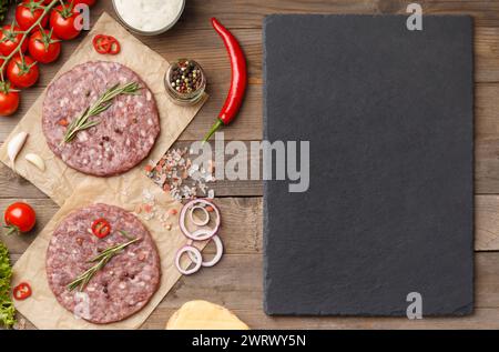 Raw burger patties with spices and rosemary, fresh vegetables, stone plate with space for text on wooden background. Stock Photo