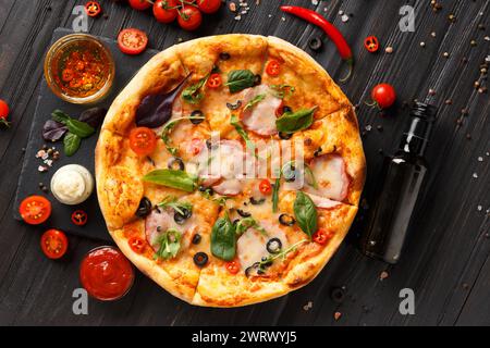Pizza with jerky meat, ham, olives, cheese and herbs, sauces and fresh vegetables on a black stone plate on a dark wooden background, top view. Stock Photo