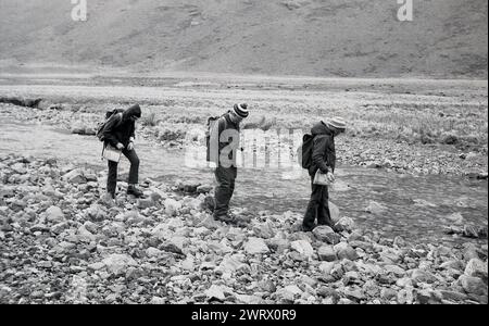1970s, two youngsters with instructor on an outward bound course, holding their maps, walking outside on rocky ground beside a shallow river in Ambleside, Lake District, Cumbria, England, UK. Stock Photo
