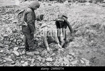 1970s, historical, three young people with accompanying adult, all wearing outdoor clothing, looking at rocks on the ground on a hillside near Ambleside in the Lake District, Cumbria, England, UK, as they take part in an outward bound course. The rocks in the Lake District include a variety of different types, including slate, sandstone, limestone and volcanic rocks. Stock Photo