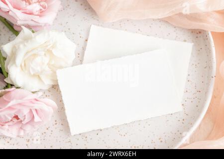 Blank cards near light pink tulle fabric and pink and cream roses  on plates top view, copy space. Wedding stationery mockup. Romantic table place wit Stock Photo