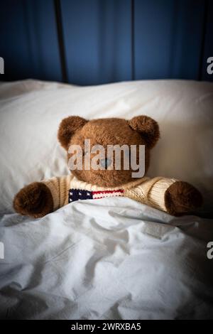 Brown teddy bear, plush toy, laying under covers in parents' bed with blue blankets and soft day light. Central view, vertical shot. Stock Photo