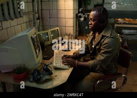 Serious African American IT specialist wearing headphones coding on old computer in abandoned building Stock Photo
