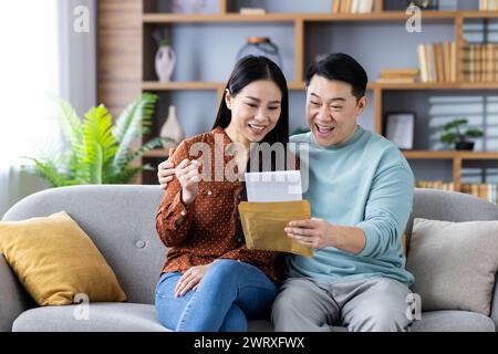 Joyful Asian couple sitting on a sofa at home, excitedly celebrating good news received in a letter. Stock Photo