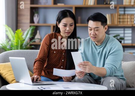 Young couple sitting on a couch at home, reviewing documents and budgeting with a laptop and papers. Stock Photo