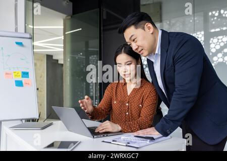 Two business professionals working together at a modern office desk with a laptop. They are discussing and pointing at the screen, engaged in teamwork and collaboration. Stock Photo