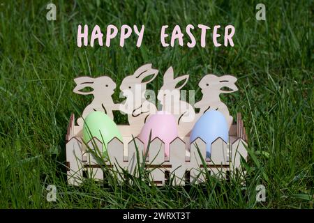 Greeting card with text Happy Easter in English. Cute wooden egg cup with painted Easter eggs standing on green grass in spring garden. Easter traditi Stock Photo