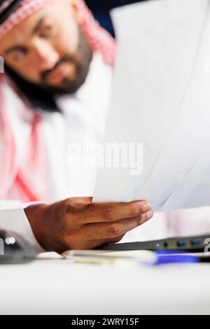 Selective focus on papers being held by a young man dressed in Arabic clothing and having a phone call. Close-up of a Muslim guy focused on examining his paperwork while speaking on a mobile device. Stock Photo