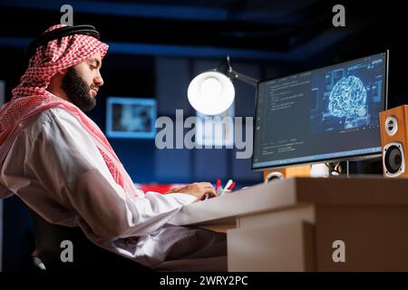 Islamic man uses AI and machine learning to ensure cyber security for a network. He monitors data centers and servers working with a PC monitor displaying advanced software and a neural network. Stock Photo