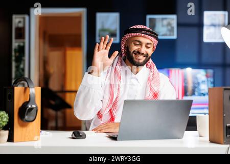 Sitting at his workplace, an Islamic guy waves at his digital laptop, starting a video conference. He is engrossed with technology, using his minicomputer to explore, interact, and conduct research. Stock Photo