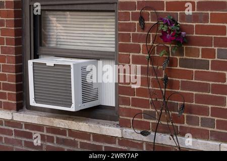 exterior view of air conditioning window unit extruding from the window sill of a red brick building with a deorative flower next to it Stock Photo