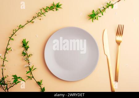 Gray plate and cutlery on light peach background with cherry blossom branches. Top view, flat lay, mockup. Stock Photo