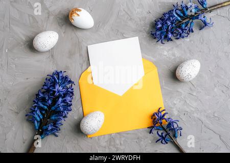 Yellow envelope with blank card, Easter eggs and blue hyacinth flowers on gray textured background. Easter concept. Top view, flat lay, mockup. Stock Photo