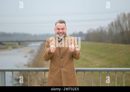 Middle aged man with gray hair and gray beard on a bridge in winter or spring giving thumbs up gesture Stock Photo