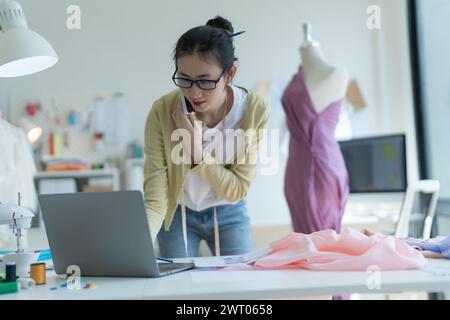 Young fashion designer wearing eyeglasses talking on the phone and checking designs on laptop computer While working on clothes sketches, stand at a t Stock Photo