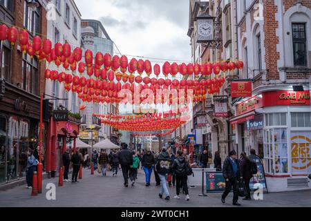 People dressed warmly walk on busy vibrant Wardour street in Chinatown London. Strings of traditional Chinese red lanterns stretch across the street. Stock Photo