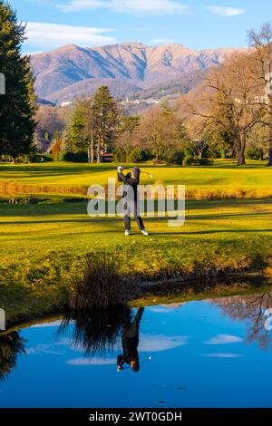 Female Golfer Reflected in a Water Pond and Hitting the Golf Ball on Fairway on Golf Course with Mountain in a Sunny Day in Switzerland Stock Photo