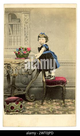 Original, charming, tinted Carte de Visite (visiting card or CDV)  of cute little Victorian girl, Victorian child, aged about 2 or 3 years old, wearing a black velvet dress, with blue triim and ribbon and a blue hairband, looking serious, standing on a velvet stool, studio of Holloway School of Photography, G Williams, Pear Tree cottage, Holloway Rd. London, U.K. Circa 1860's Stock Photo