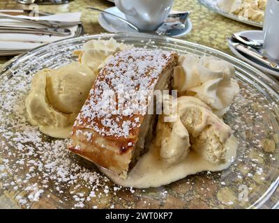 Café table with a glass plate with delicious Apfelstrudel with icecream and whipped cream outdoors in summer. Stock Photo