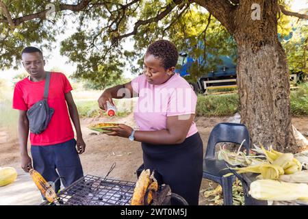 african woman street vendor selling grilled corn to a man client on a street in the city on the sidewalk, car in the background Stock Photo