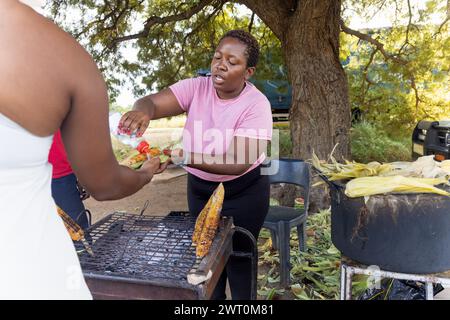 african woman street vendor selling grilled corn to a  client on a street in the city on the sidewalk, pot boiling in the background Stock Photo