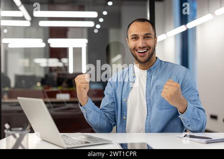 Portrait of a happy and satisfied young Muslim man sitting in the office at the table with a laptop and rejoicing in success, looking at the camera and showing a victory gesture with two hands. Stock Photo