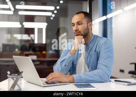 Pensive young hispanic man working and studying in office, sitting at desk holding head in hand and looking at laptop, analyzing data, looking for solution, thinking about project. Stock Photo